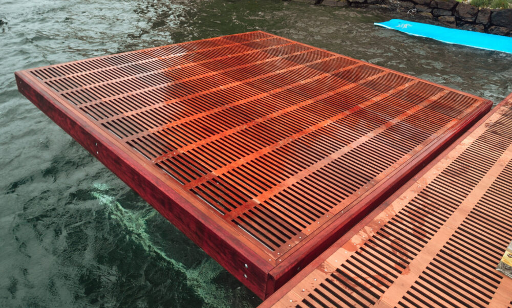 A large, rectangular dock of reddish-brown wood, perfect for Custom Boat Lifts, floats by the stone shoreline, adjacent to another structure and a blue floating mat. Evenly spaced slats enhance its design and durability on the water.