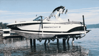 A white and black MasterCraft speedboat is lifted out of the water by a premium boat lift on a dock, tilting slightly as it rises above the lake surface under a clear sky.