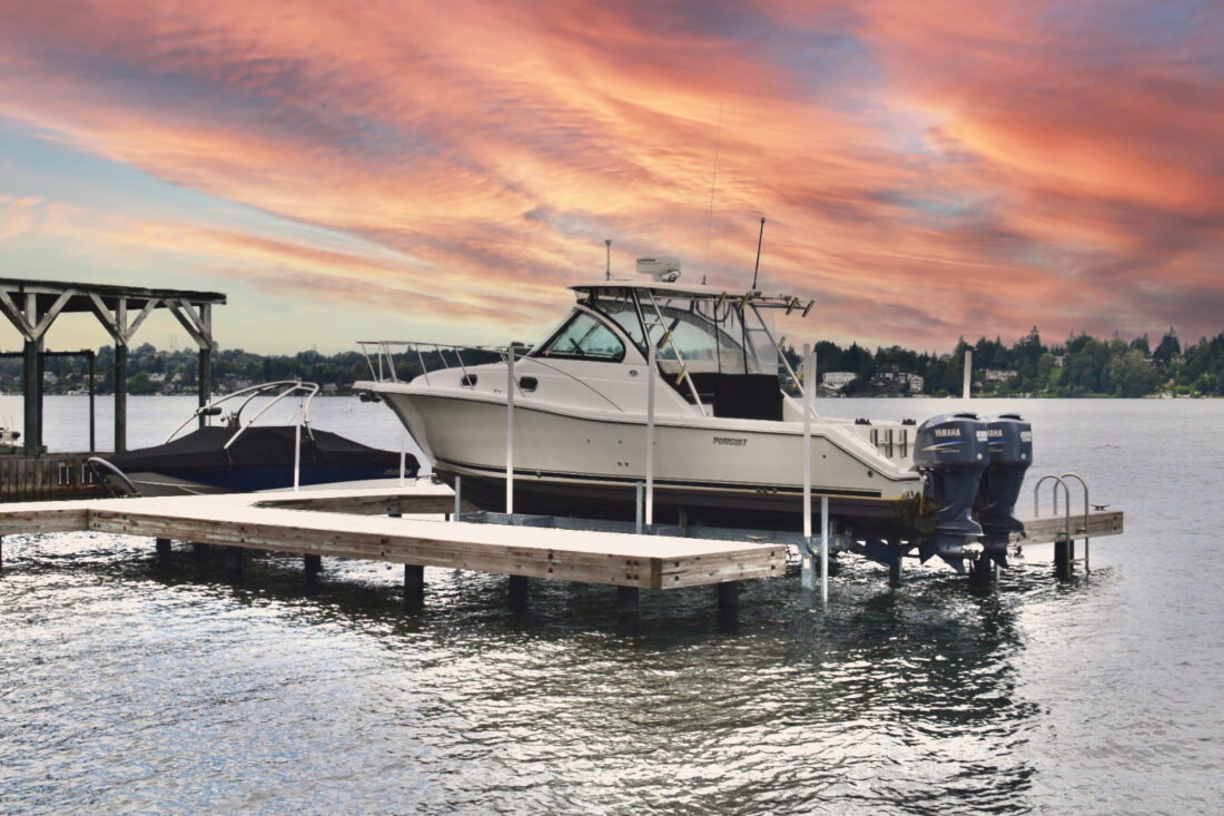 A white motorboat is docked on a lift beside a smaller boat, unveiling the power of Basta Boatlifts, with a vibrant sunset sky and calm water in the background. Trees and houses are visible along the distant shoreline.