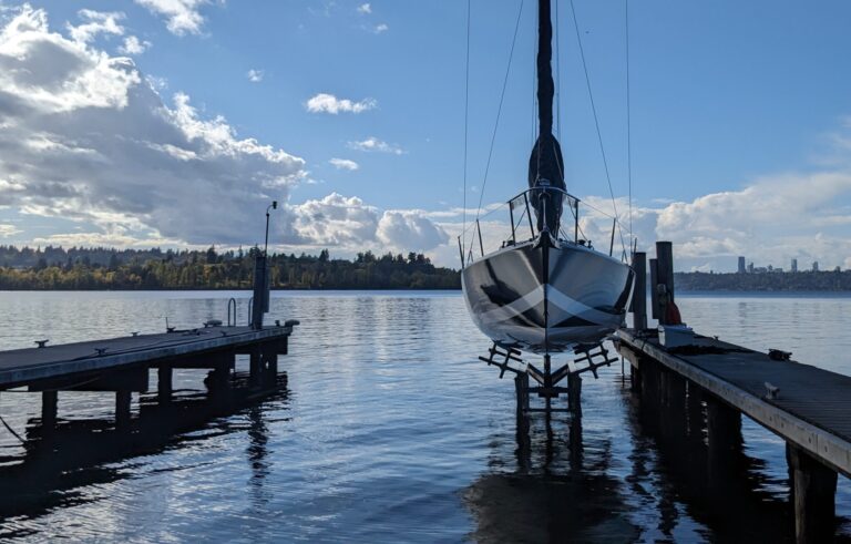 A sailboat rests on a sailboat lift above the water between two wooden docks on a calm lake, with trees and distant city buildings visible under a partly cloudy sky.