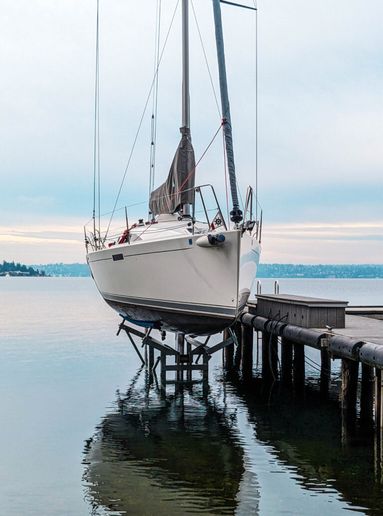 A white sailboat rests on a sailboat lift above calm water next to a dock, with a serene lake and distant shoreline stretching under a cloudy sky.