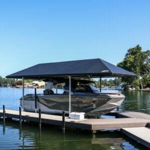 A sleek gray motorboat is parked under a boat lift canopy on a wooden dock by a calm lake, surrounded by trees and houses, with a clear blue sky overhead.