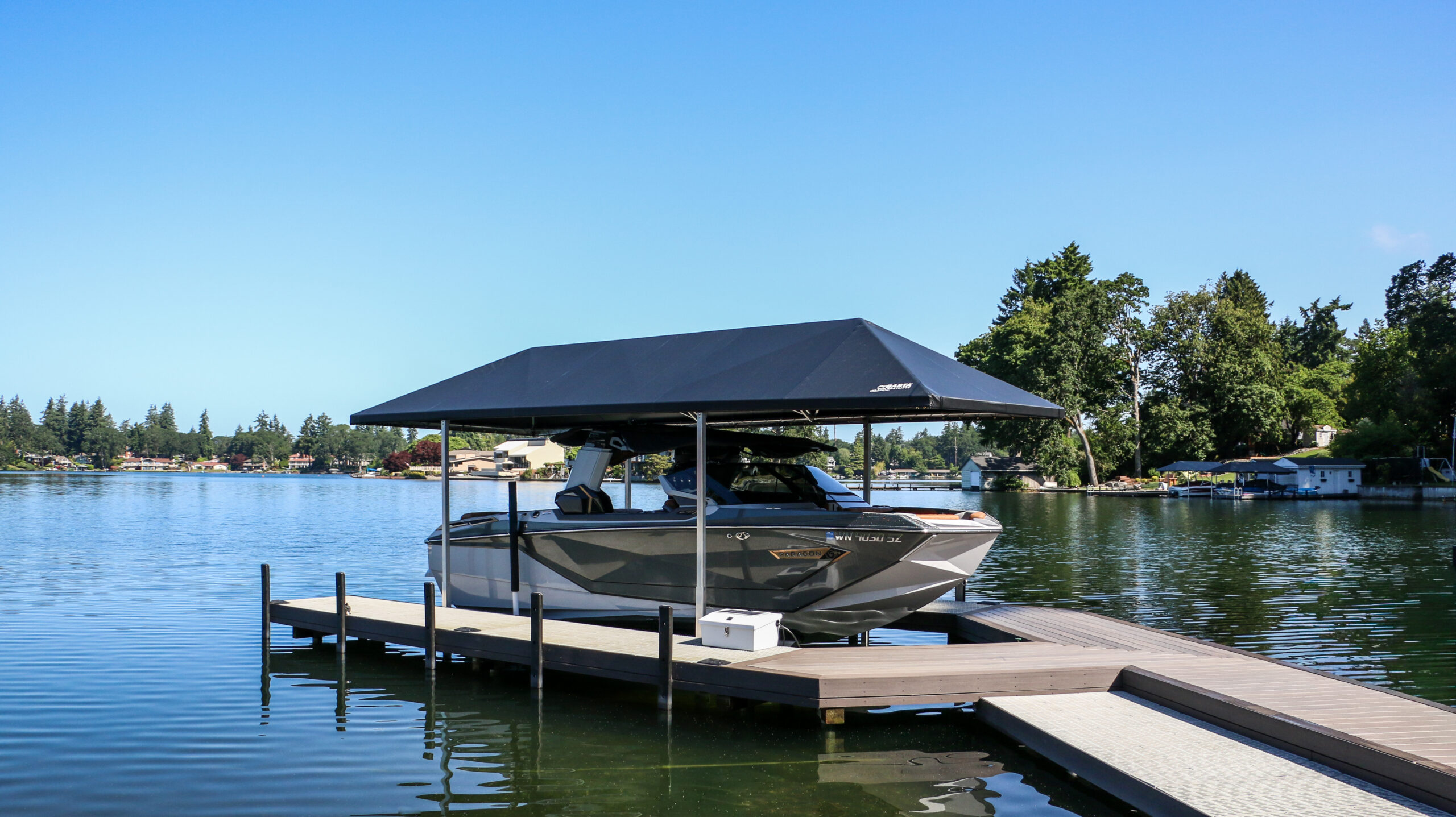 A sleek gray motorboat is parked under a boat lift canopy on a wooden dock by a calm lake, surrounded by trees and houses, with a clear blue sky overhead.