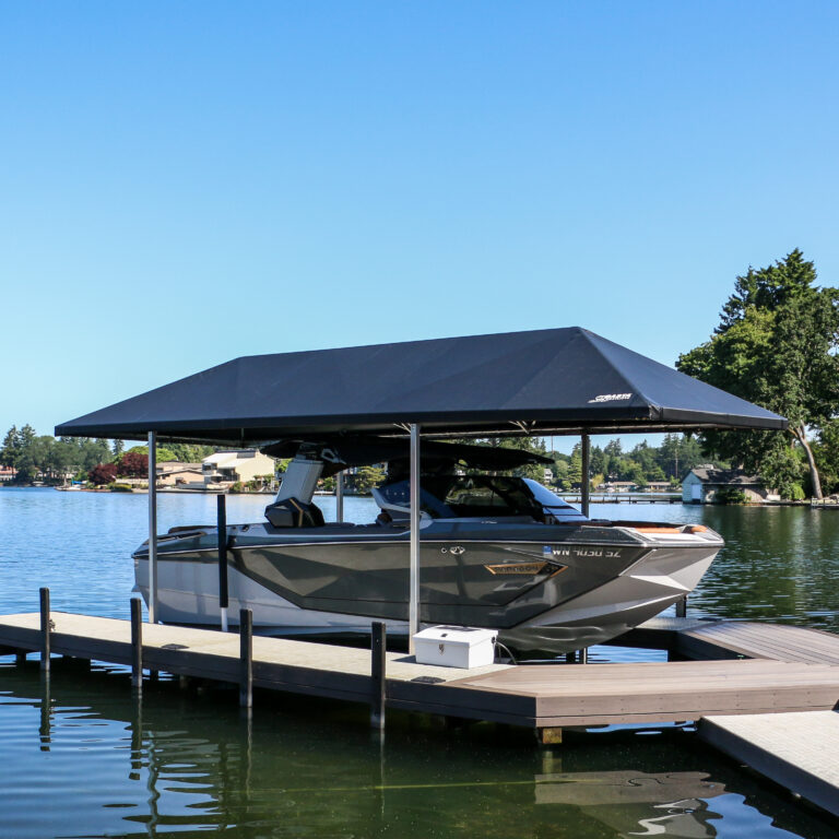 A sleek gray motorboat rests under a black boat lift canopy on a lake, surrounded by a wooden pier. Trees and houses frame the scene beneath a clear blue sky.